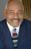 Thom Gossom Jr. wearing a suit and tie smiling at the camera
