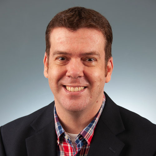 A man in a blazer and plaid shirt smiling against a neutral background at Auburn University at Montgomery.