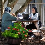 Two students work on their laptops and eat lunch outside on a picnic bench