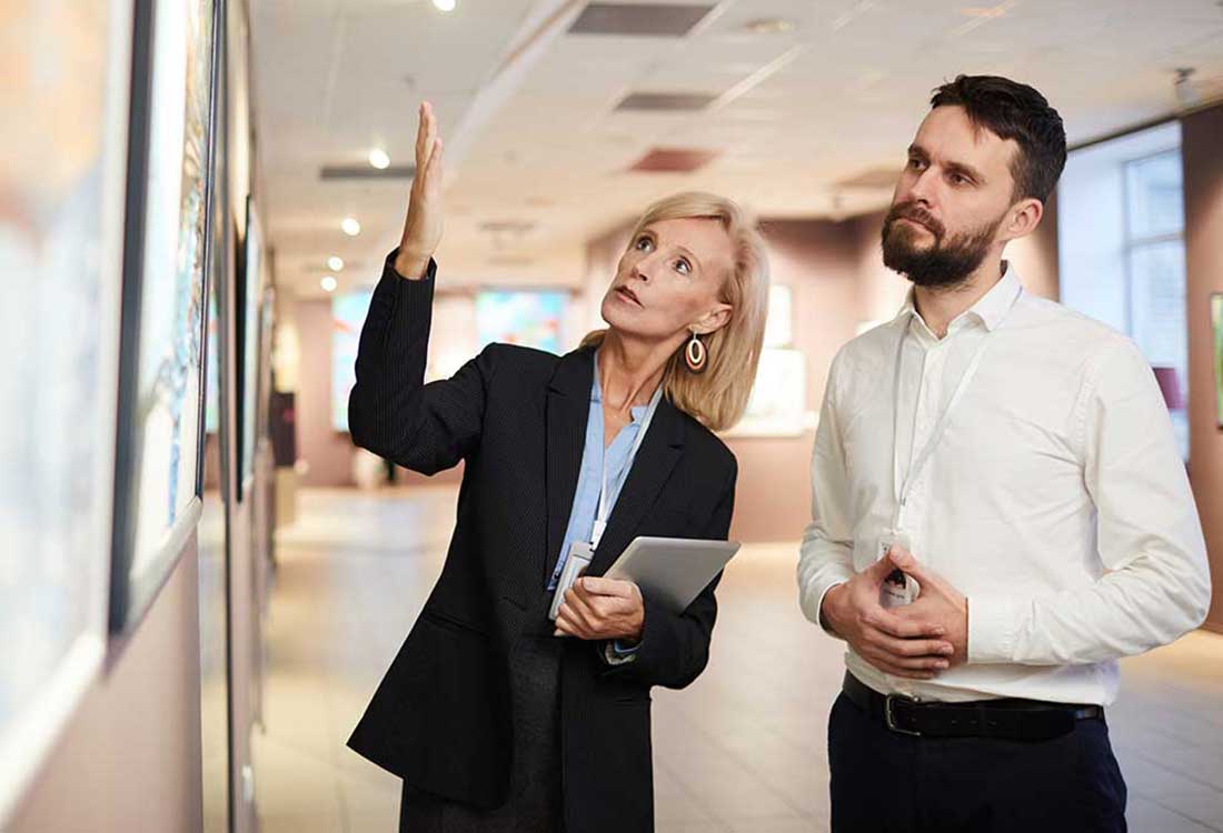 A woman guides a man through an Auburn University at Montgomery art gallery, discussing a painting on display.