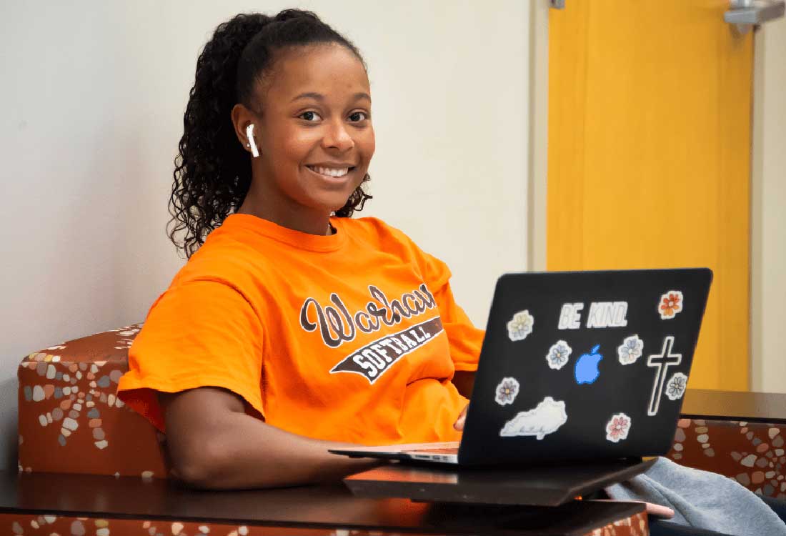 Auburn University at Montgomery student in orange Warhawks softball shirt studying on a laptop in a campus lounge