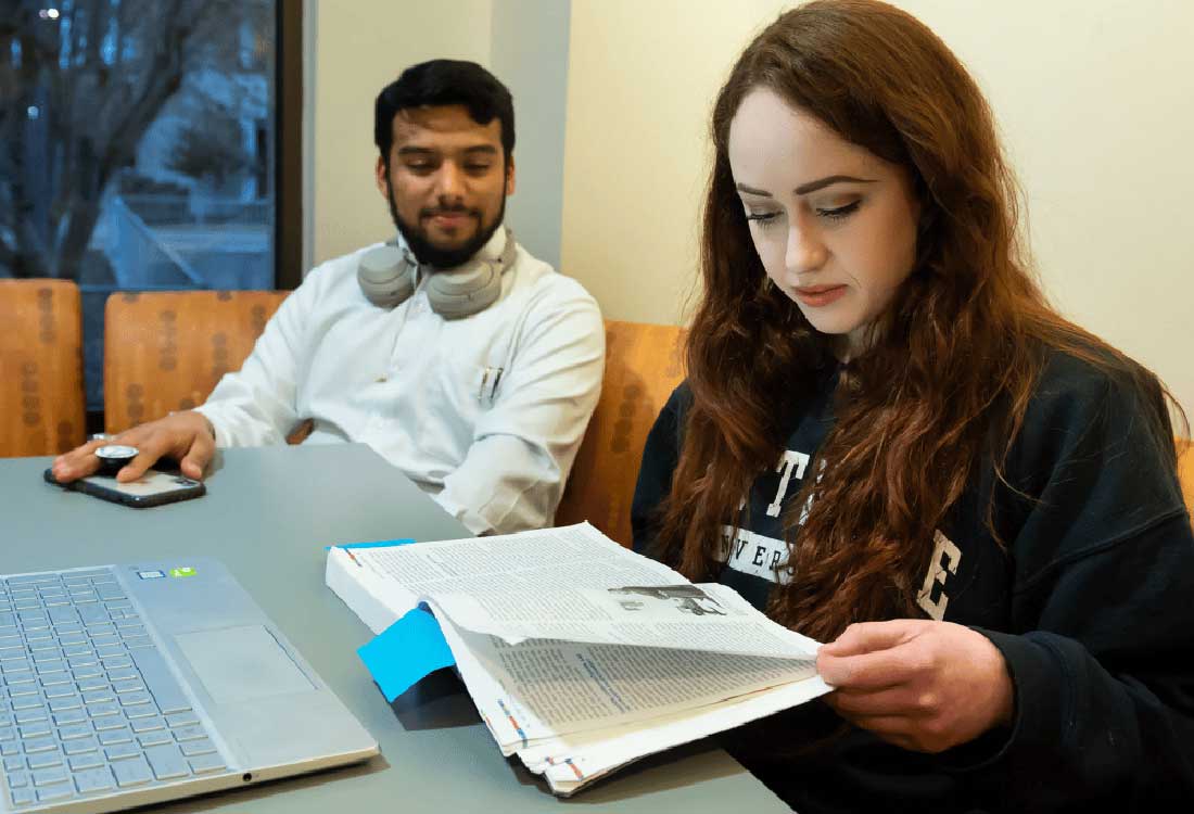 AUM students studying together in campus library; woman reading textbook, man with laptop and headphones at table.