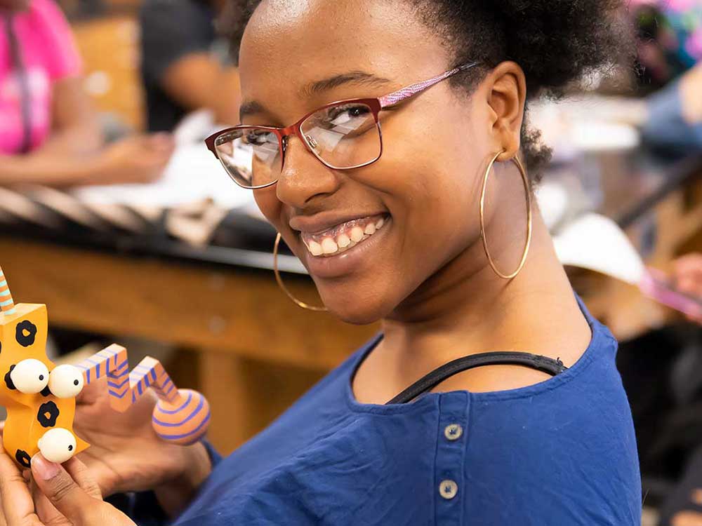 A smiling woman in glasses holds a colorful, playful craft with eyes at a classroom setting.