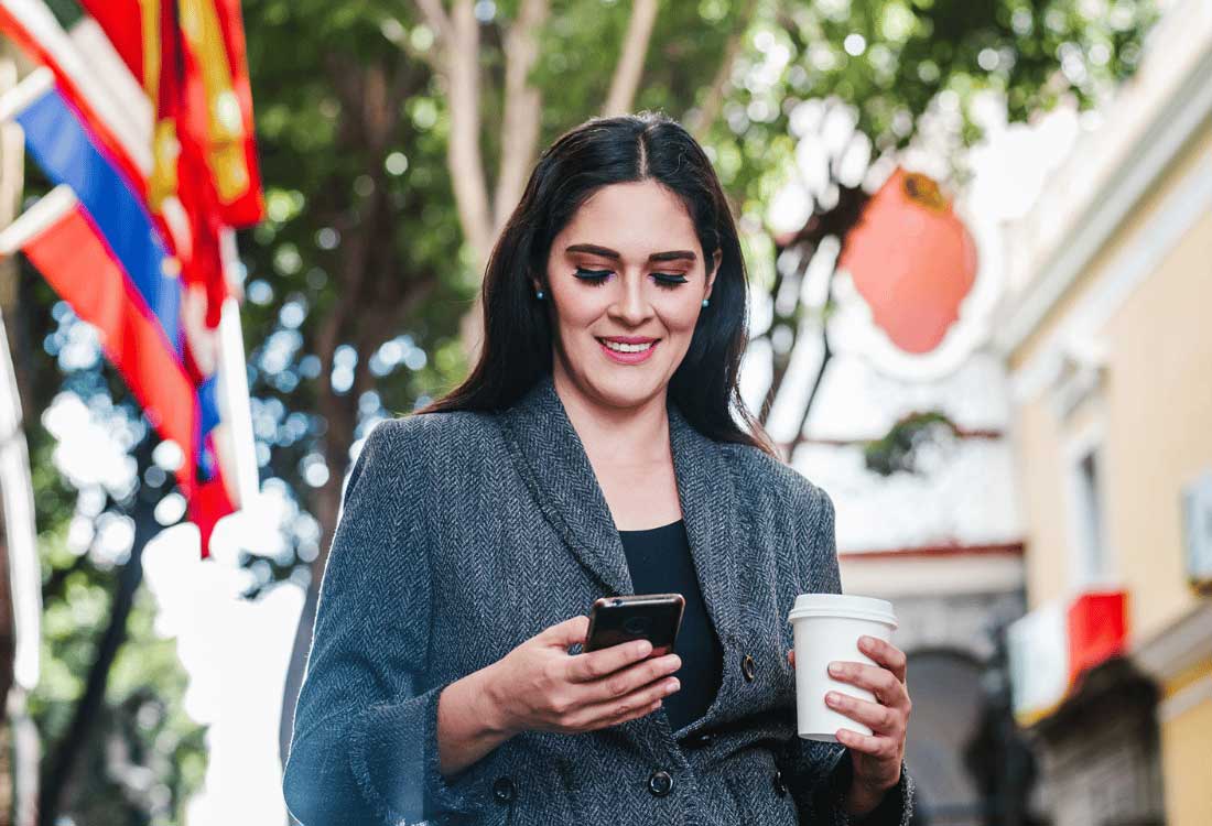 Auburn University at Montgomery student in business attire checks phone with coffee on campus.