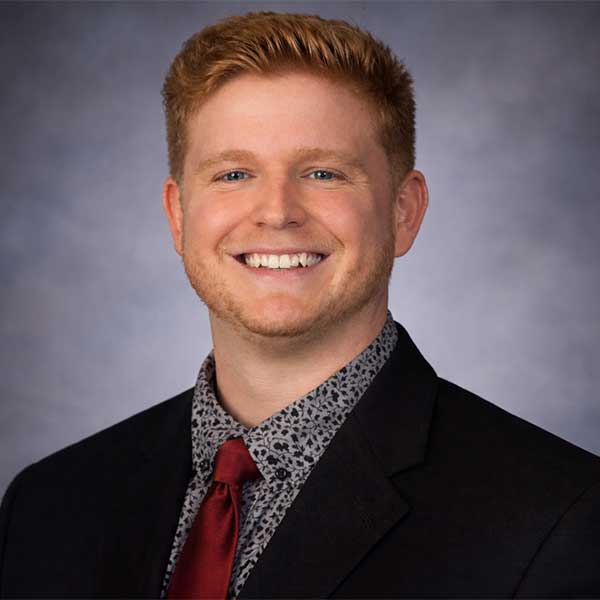 A person in a suit smiling with a gray and red tie against a gray background.