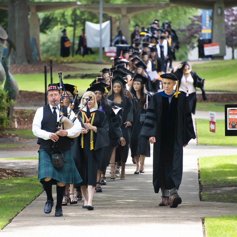 A processional of college students wearing black caps and gowns walks toward the Auburn University at Montgomery Athletics Complex.