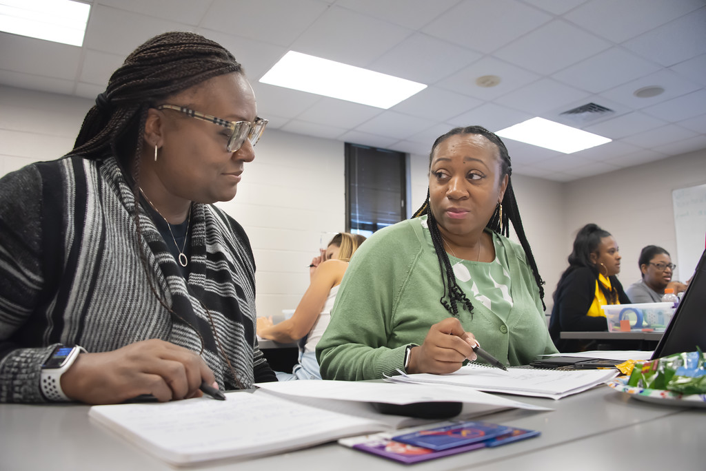 Two female educators discuss notes while seated at a table during a workshop on the Auburn University at Montgomery campus.