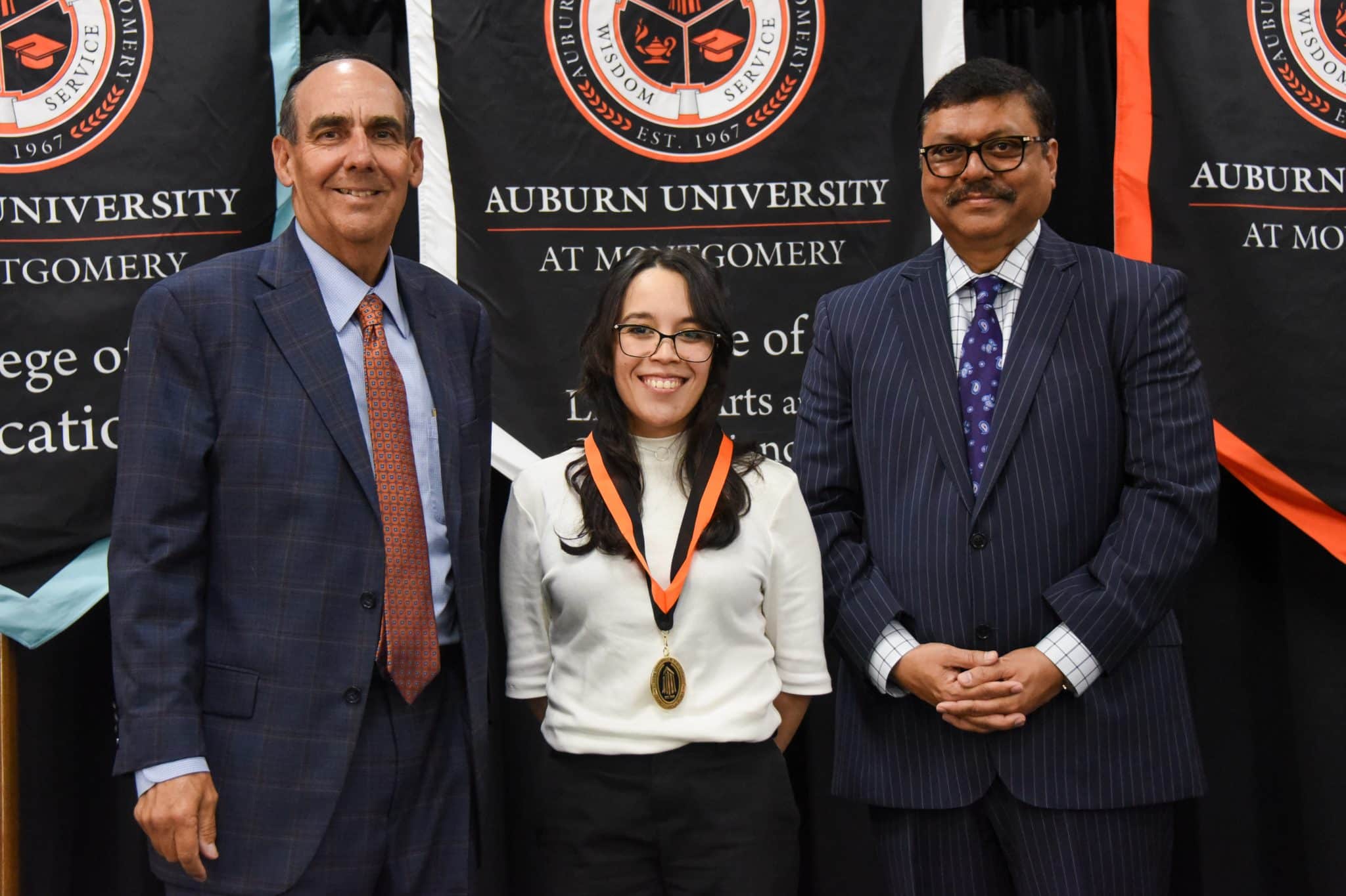 Three people stand smiling at an Auburn University at Montgomery event with banners in the background.