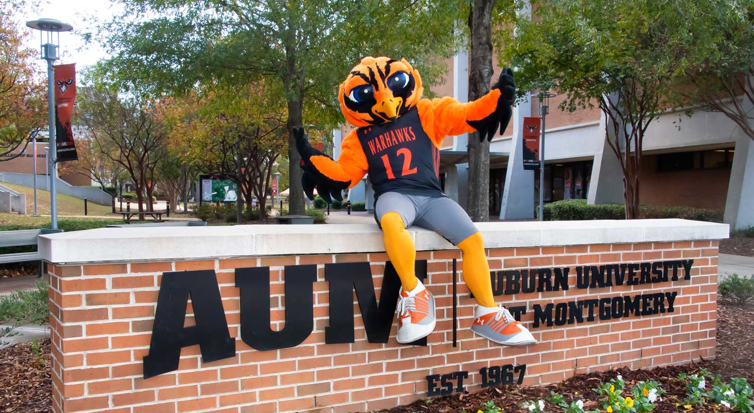 AUM mascot sitting on Auburn University at Montgomery sign with trees and campus in the background.