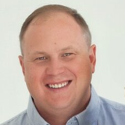 A Person Smiling In A Blue Collared Shirt Against A White Background.