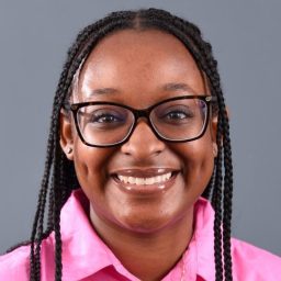 A Person Smiling, Wearing Glasses And A Pink Shirt, With Braided Hair, Against A Gray Background.