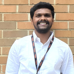 A Person Smiling In A White Shirt With An Auburn University At Montgomery Lanyard, Standing Against A Brick Wall.
