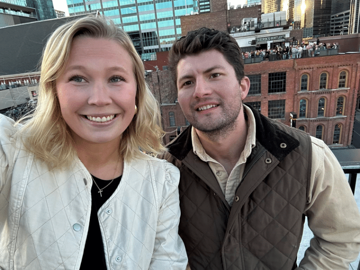 A woman and a man smiling on a rooftop with city buildings in the background at sunset.