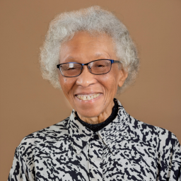 An Elderly Woman With Glasses Smiles In A Patterned Shirt Against A Plain Background.