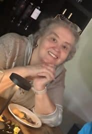 A woman smiling at a dining table with a plate of food in a cozy setting.