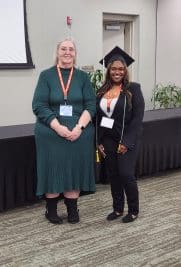 Two women smiling, one in a graduation cap and gown, standing in a conference room at Auburn University at Montgomery.