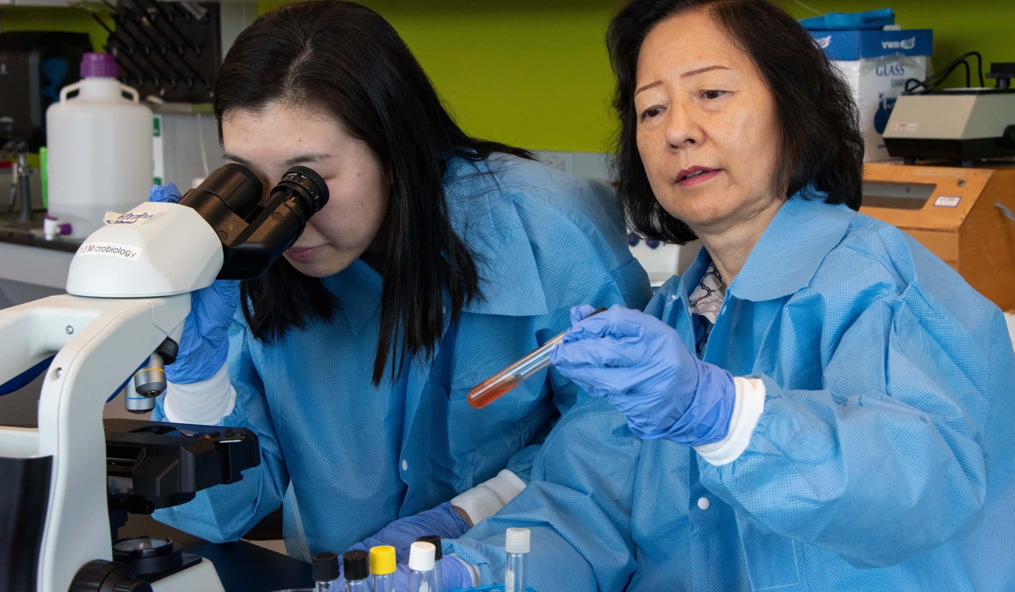 Two scientists in blue lab coats examine a test tube and a microscope.