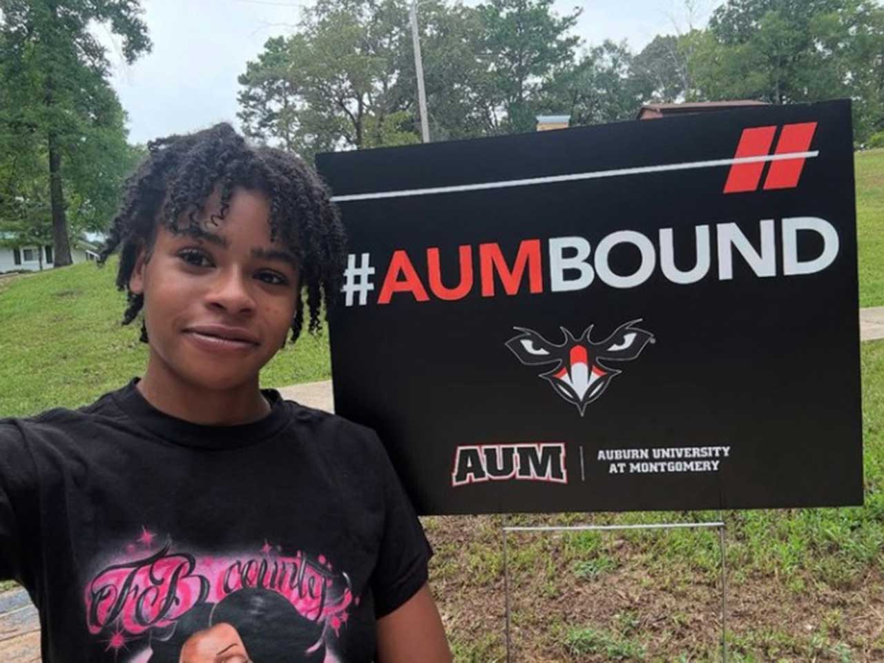 A person smiling next to an Auburn University at Montgomery #AUMBbound sign on a grassy area.