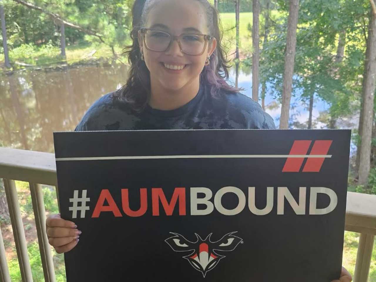 A woman smiling, holding a sign with #AUMBOUND, standing outside with trees and a pond in the background.