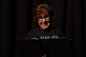 A Woman Wearing Glasses And A Theatre AUM Shirt Stands In Front Of A Lectern On A Dark Stage.