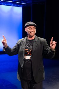 A Man Wearing A Black Hat And Jacket Gestures On Stage Under Blue Lighting At A Theater Event.