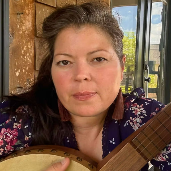 A woman holding a banjo, sitting in a wooden interior with a window view.
