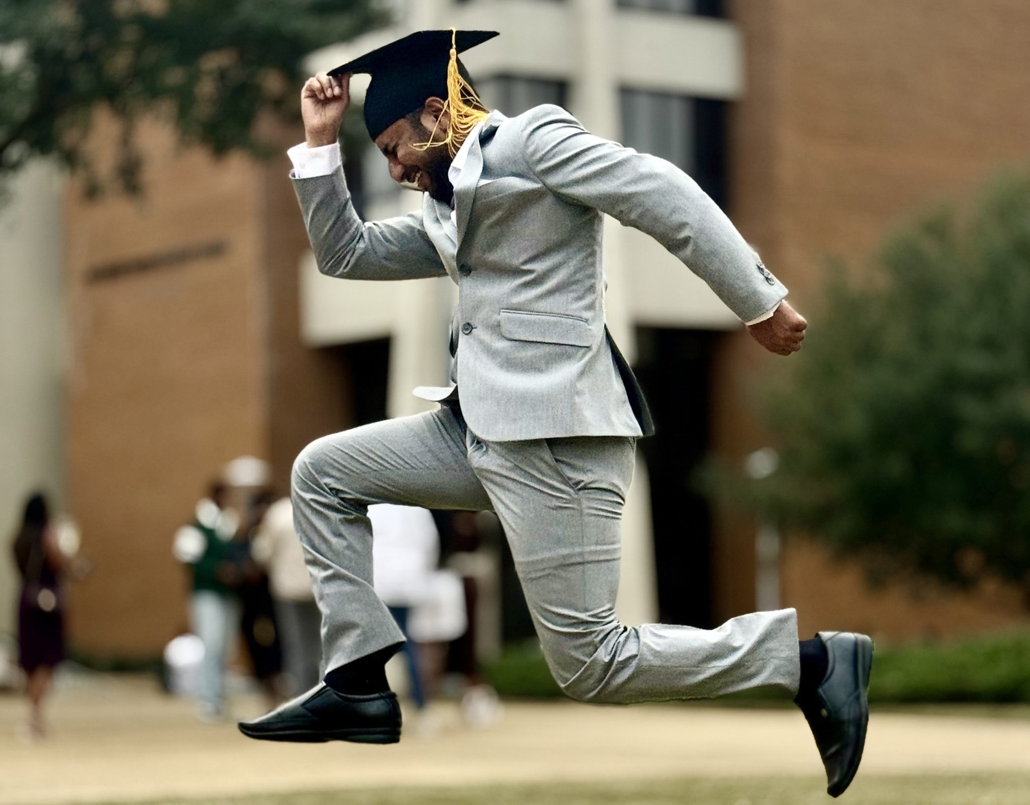 Graduate in gray suit jumps joyfully with cap at Auburn University at Montgomery campus.