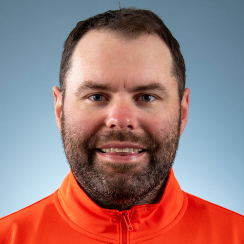 A person in an orange shirt smiling at the camera against a blue background.