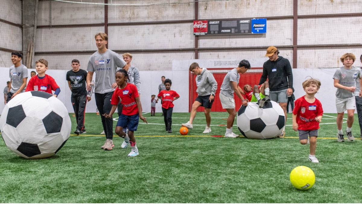 A group of children and student-athletes playing soccer indoors on a green turf field.