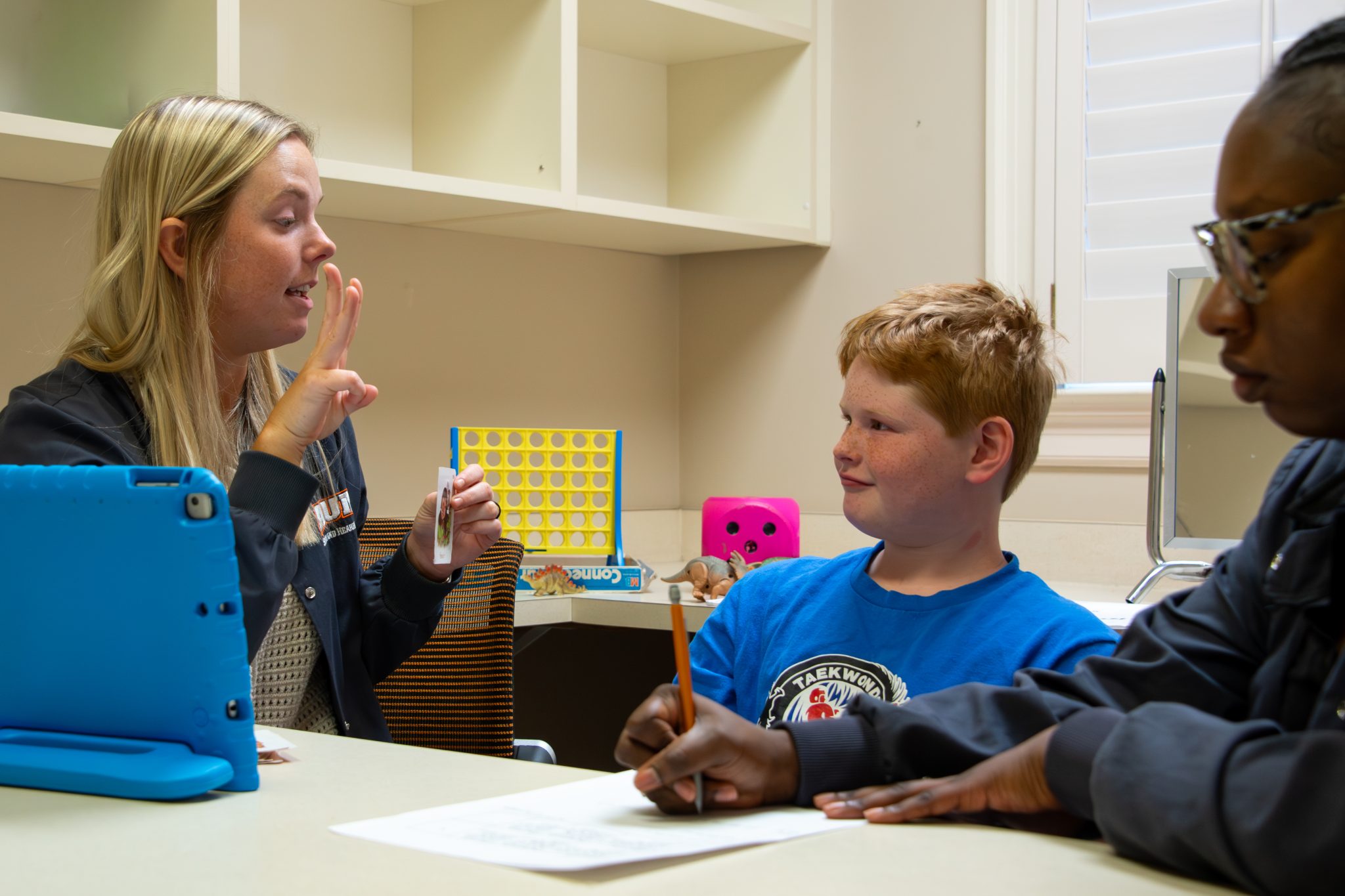 Auburn University at Montgomery speech therapy session: student clinician guides a child with flashcards as another student takes notes.