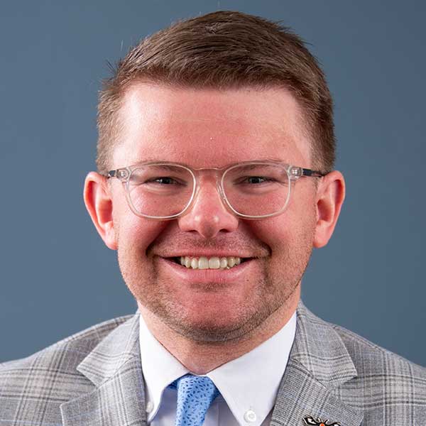 Auburn University at Montgomery portrait of a man in glasses, gray suit and blue tie, smiling against a blue backdrop