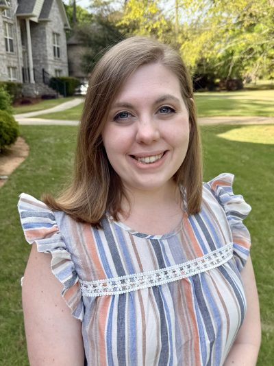 A woman smiling outdoors on the Auburn University at Montgomery campus, standing on a sunny lawn near campus buildings.