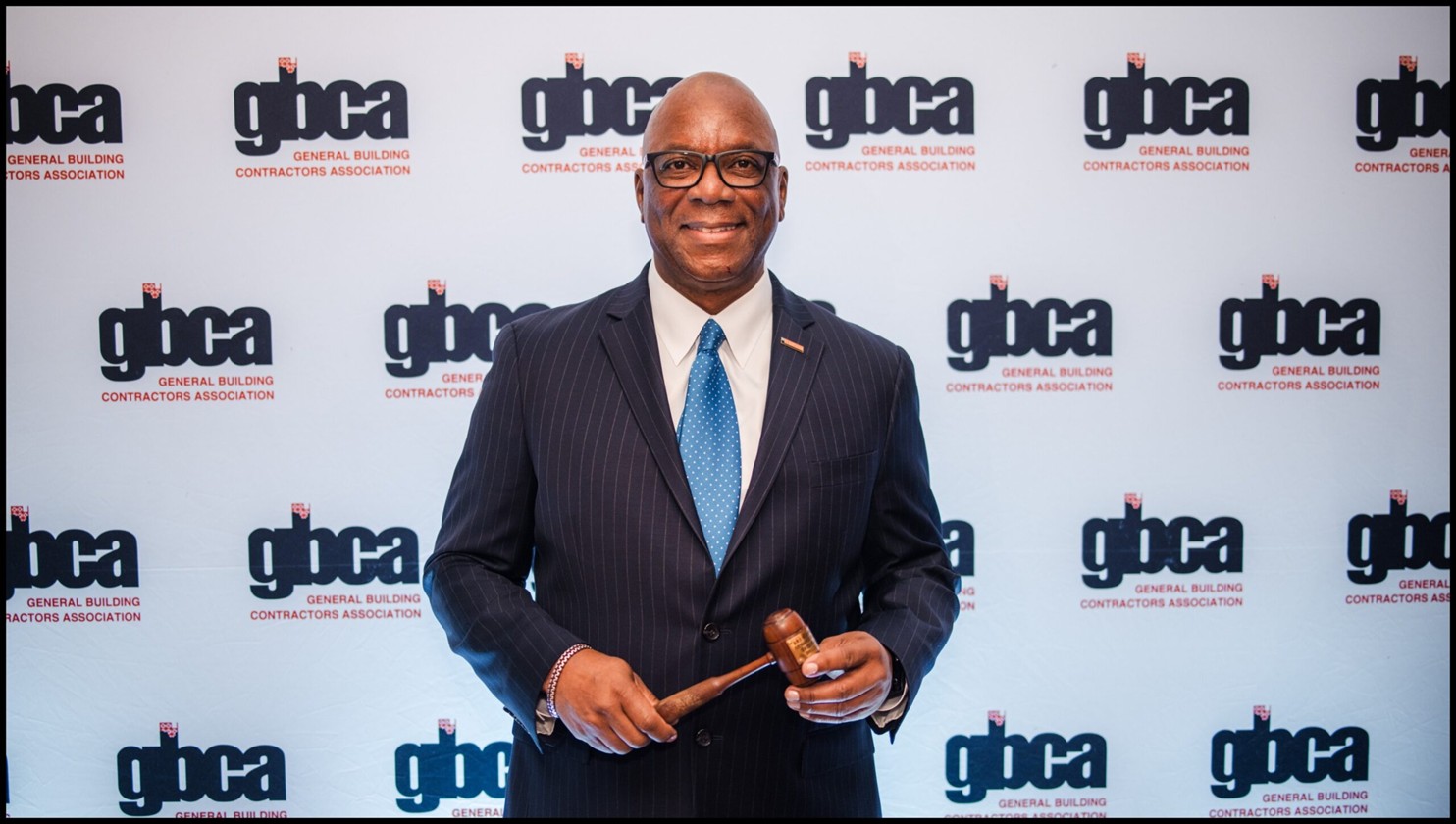 Angelo Perryman smiling and holding a gavel at a GBCA event backdrop.