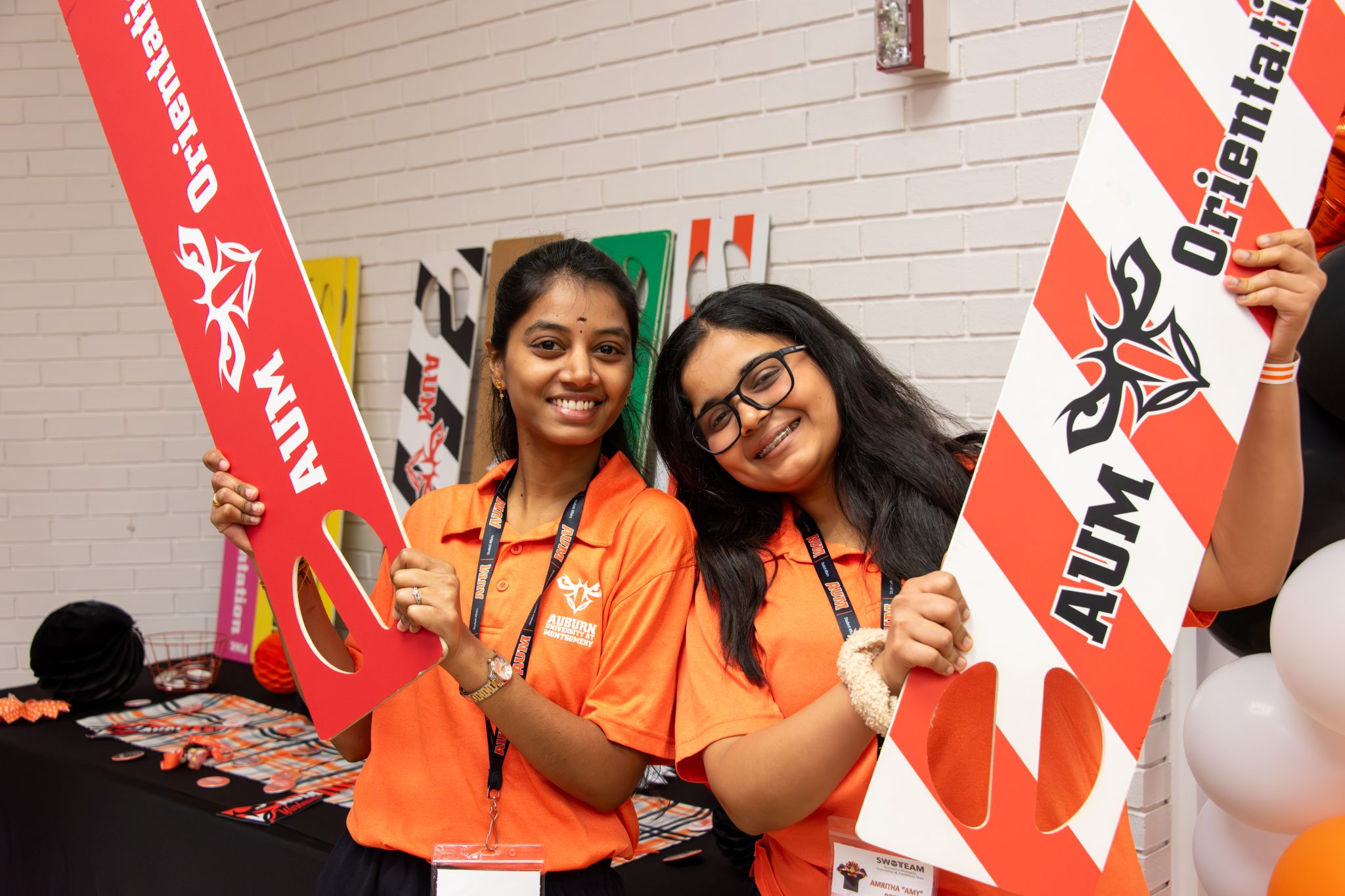Two AUM students at Auburn University at Montgomery hold AUM Orientation signs during campus event, smiling in orange polos.