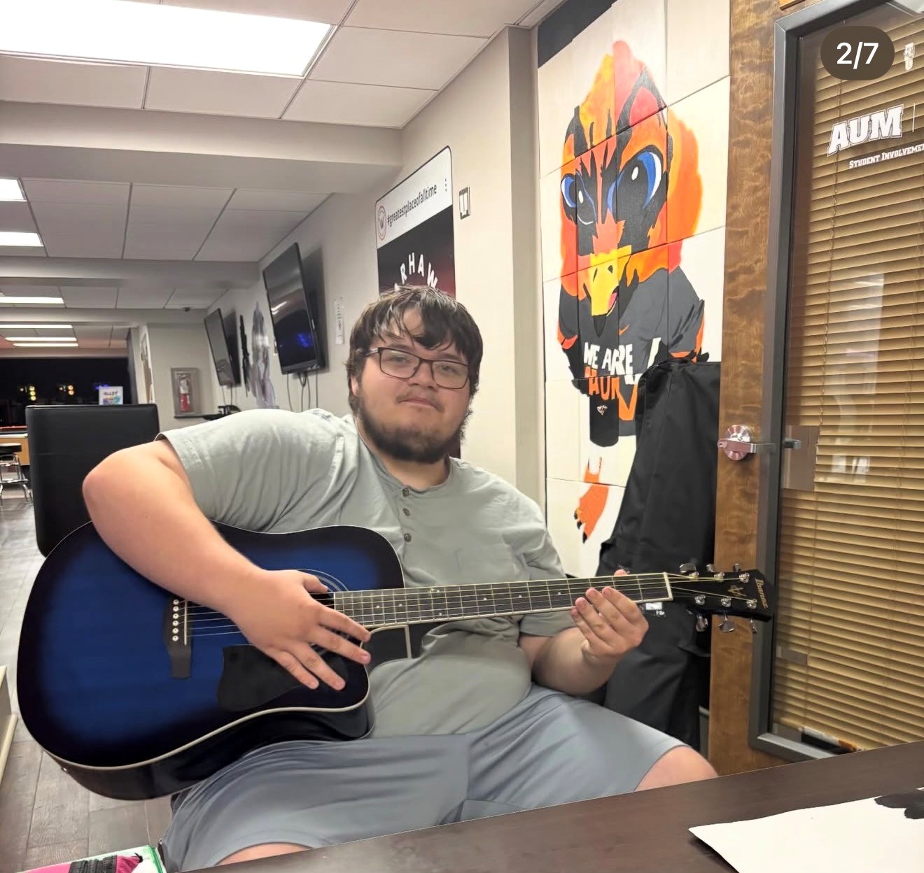 AUM student plays blue acoustic guitar in student center by Warhawk mural at Auburn University at Montgomery.