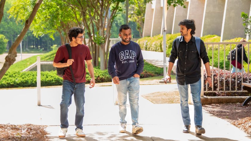 Three Auburn University At Montgomery Students Walking Together On A Sunny Campus Path With Backpacks.