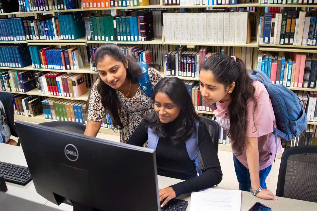 Auburn University at Montgomery students collaborating at a library computer, studying with books and notes.