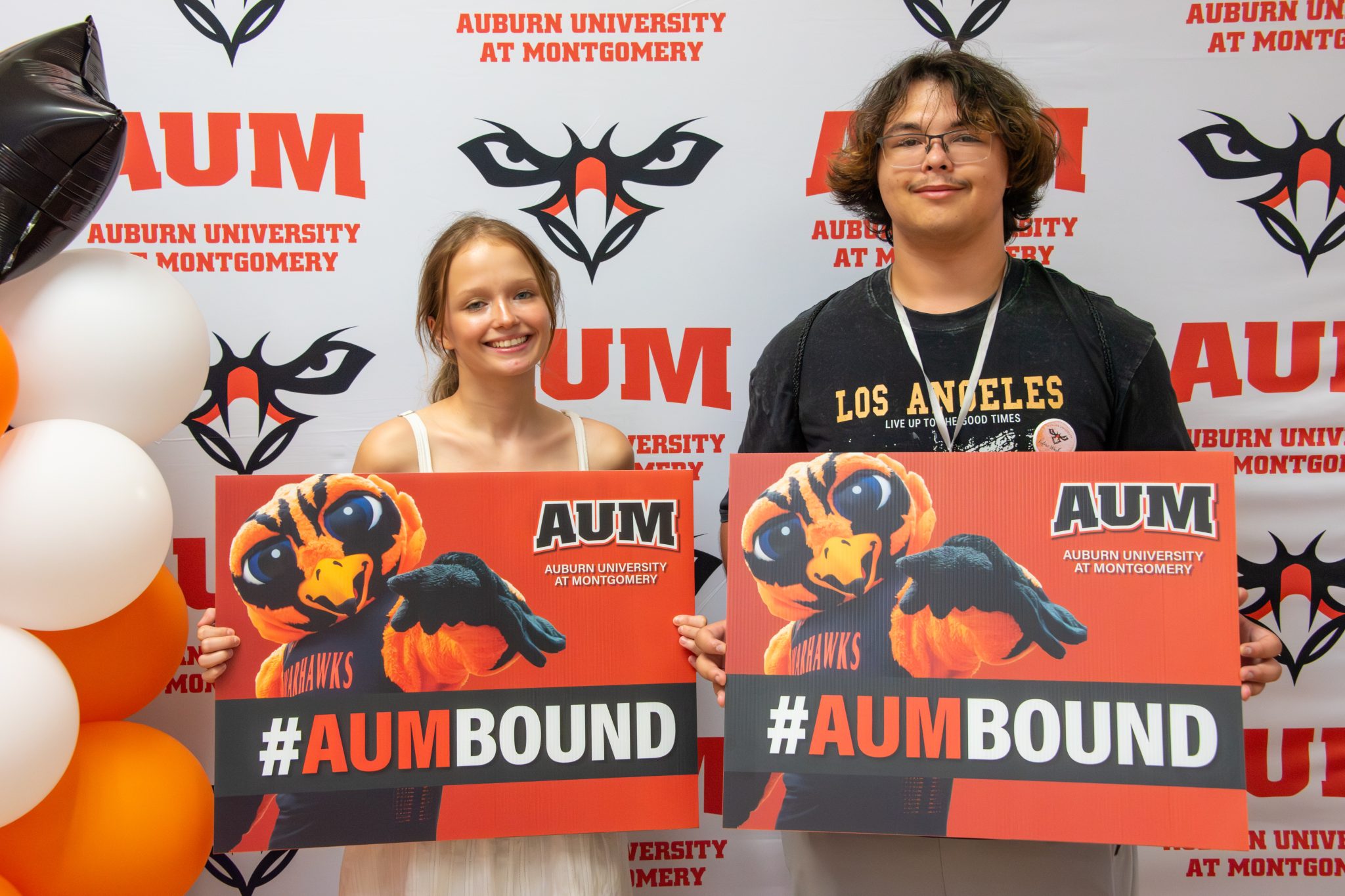 Two students at Auburn University at Montgomery hold #AUMBOUND Warhawks signs by balloons and AUM backdrop.