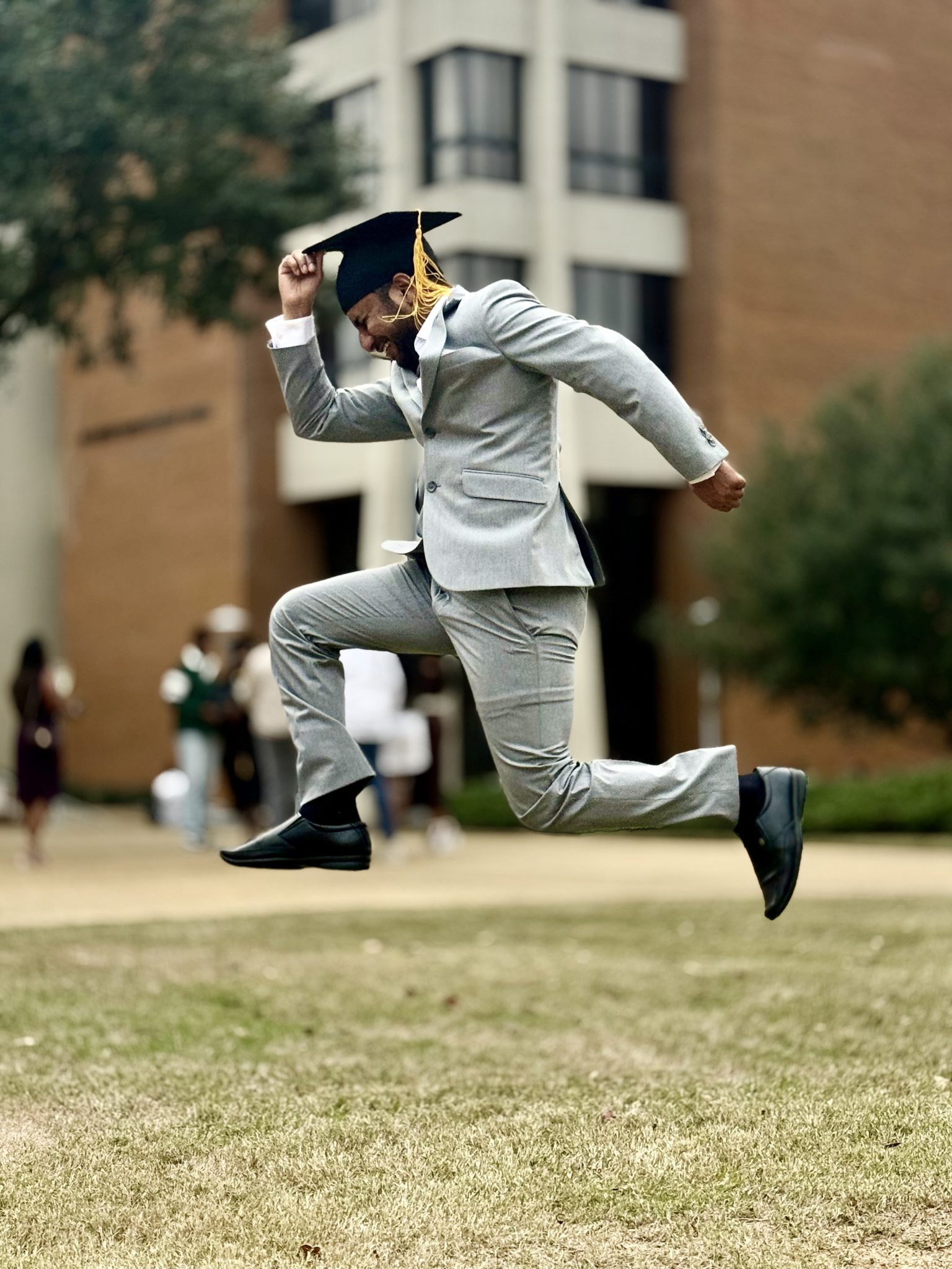 AUM graduate, Ravikanth Gwoda, in gray suit jumping with cap on campus lawn, celebrating commencement.