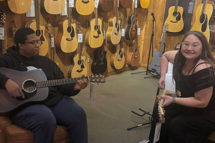 Auburn University at Montgomery students play acoustic guitars in a shop with a wall of guitars.