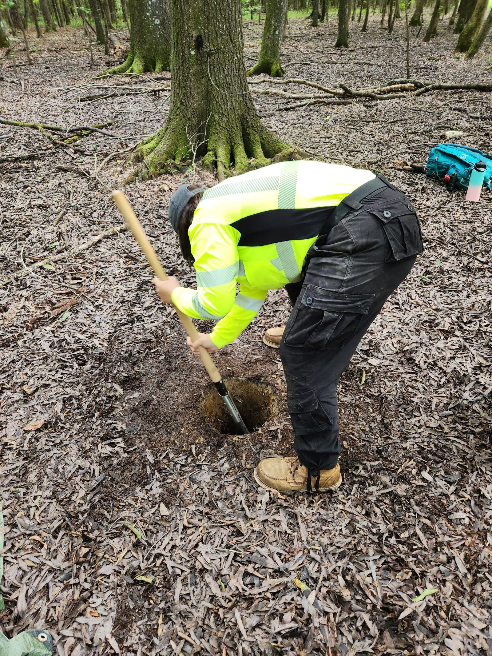 A person in high-visibility jacket digs soil sample pit in forest during Auburn University at Montgomery field research.