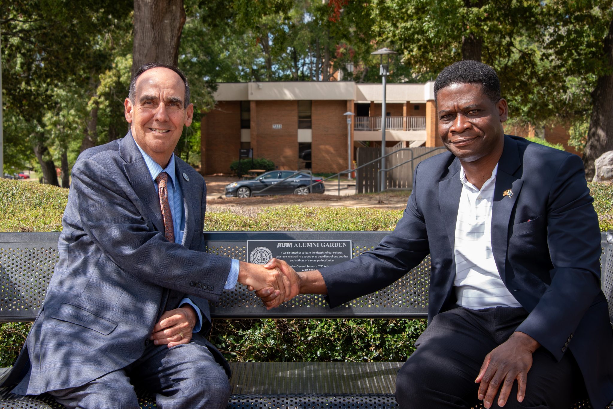Two men in suits shake hands on an AUM Alumni Garden bench at Auburn University at Montgomery campus.