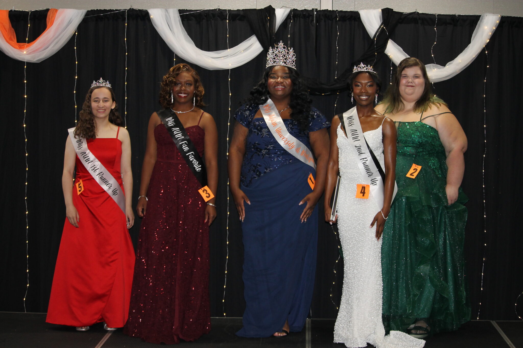 Auburn University at Montgomery pageant contestants pose on stage in evening gowns and sashes, as the winner wears a crown.